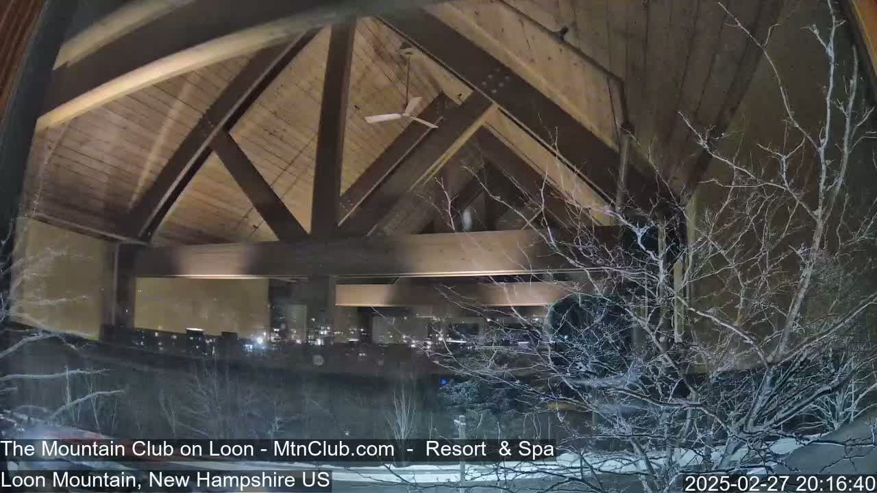 A view through a window shows a snow-covered landscape at night, with a wooden beamed ceiling and interior lights visible inside the building.