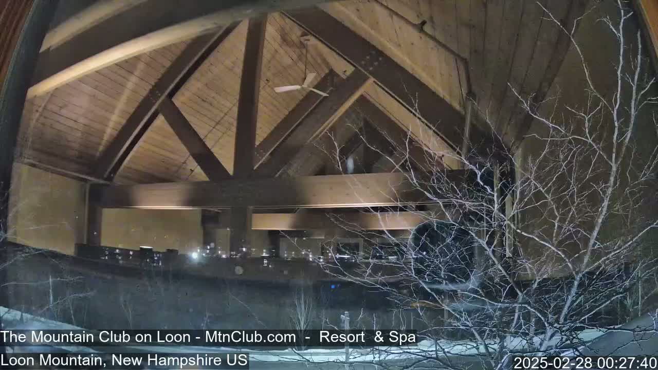 A nighttime view from inside a building with a wooden, vaulted ceiling shows a snow-covered exterior through a window.
