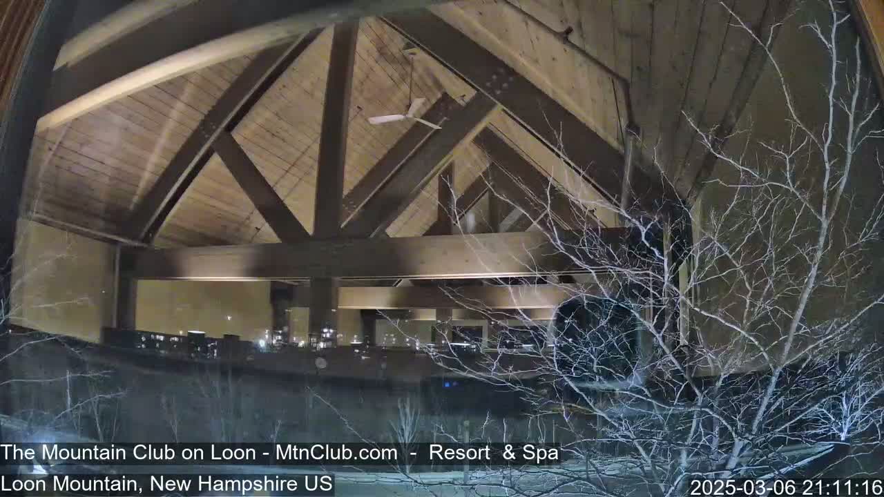 A nighttime view through a window shows a snow-covered landscape and the interior of a building with a wooden, vaulted ceiling and lighting.