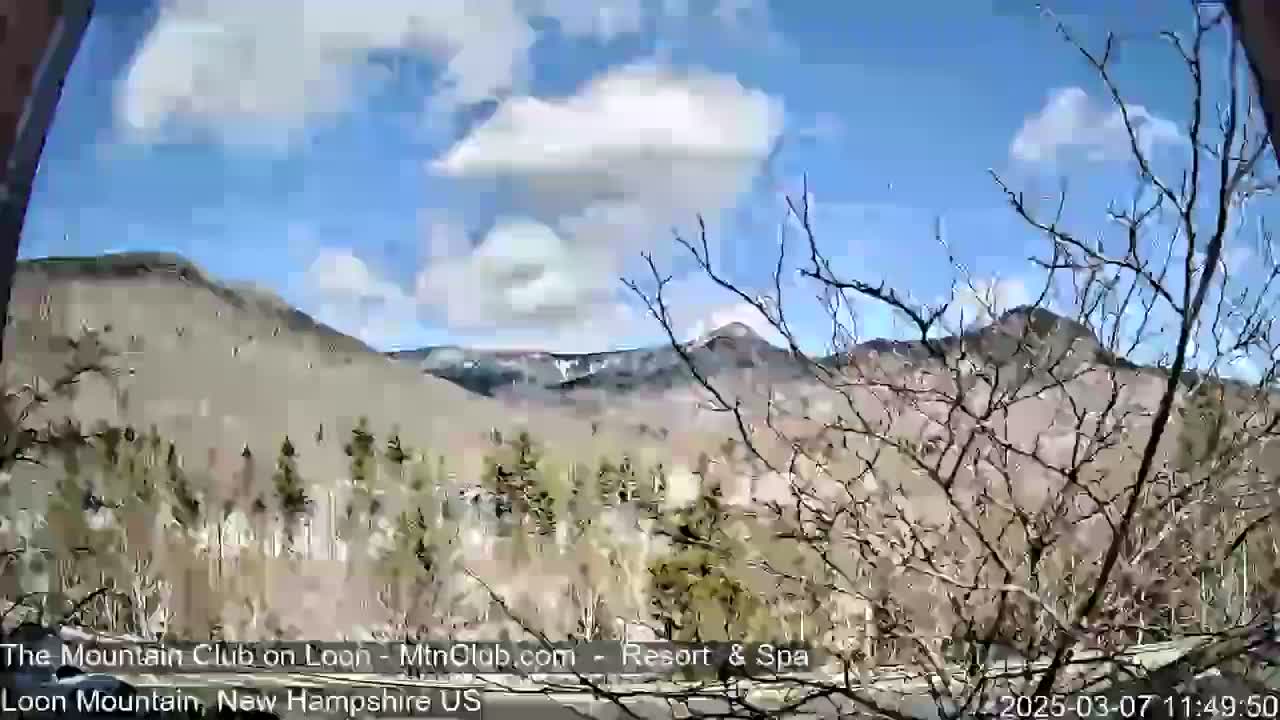 A partly cloudy blue sky overlooks a mountainous landscape with leafless trees in the foreground.
