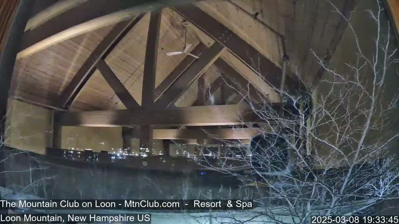 A view through a window shows a building's interior with a high, wood-beamed ceiling, a ceiling fan, and a snowy, nighttime exterior.