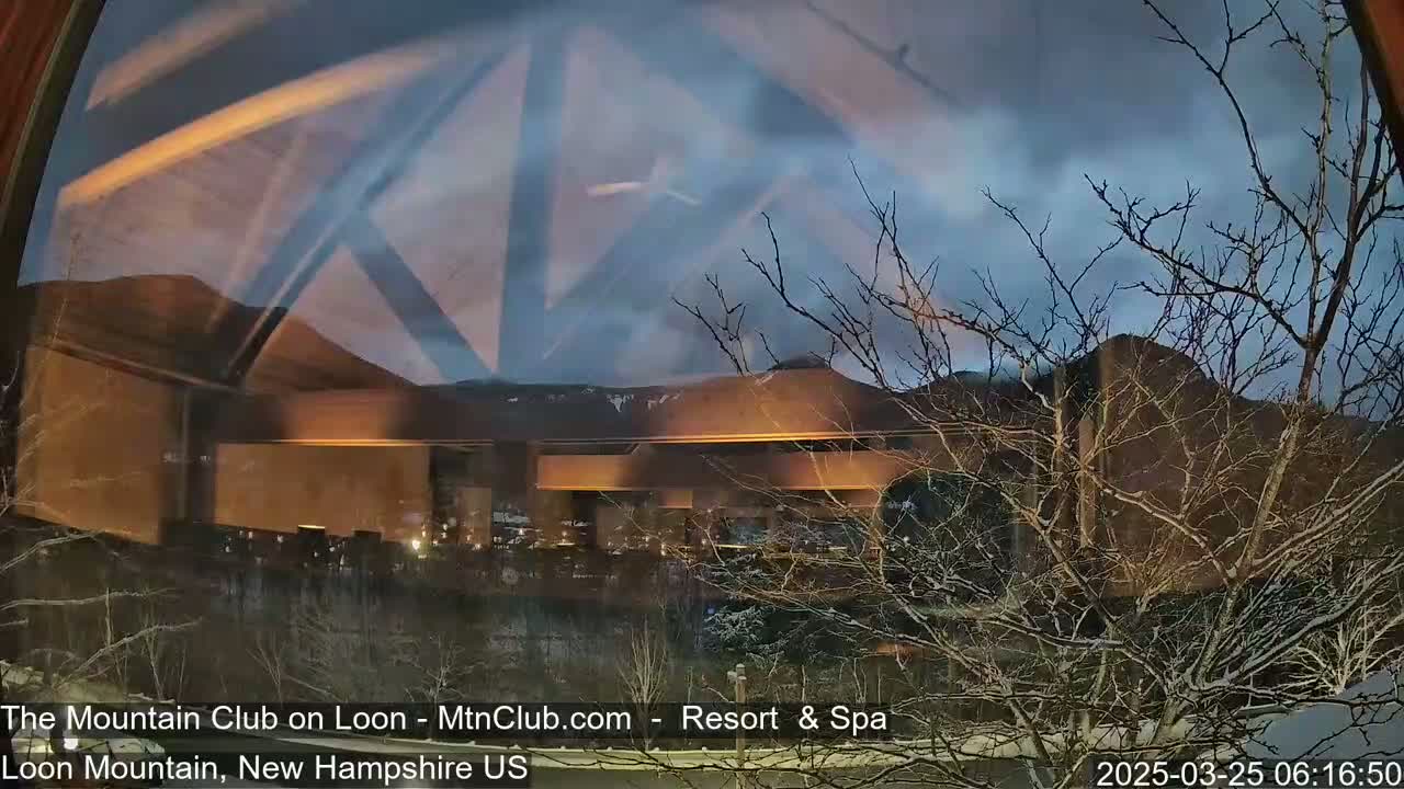 A view through a window shows a snow-dusted landscape at twilight, with bare trees in the foreground and a large building illuminated in the midground against a backdrop of mountains under a cloudy sky.