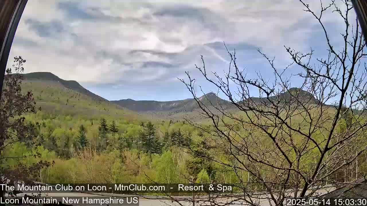 A partly cloudy sky overlooks a mountainous landscape with lush green trees in the foreground.