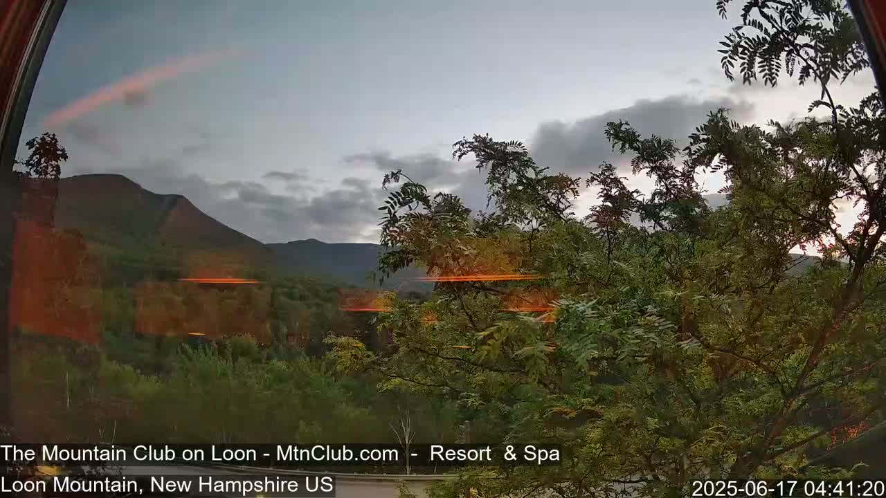 A view through a curved window shows a partly cloudy sky over a mountain range and a foreground of green trees.