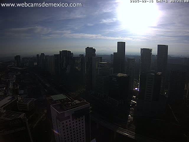 A partly cloudy, sunlit cityscape of numerous high-rise buildings is viewed from an elevated perspective.