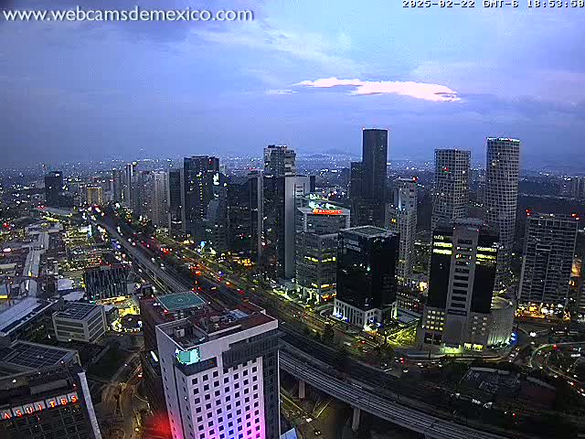A nighttime aerial view of a city with many tall buildings and a highway, under a mostly clear sky with some clouds.