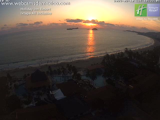A beach resort at sunset overlooks a calm ocean with a setting sun reflecting on the water.