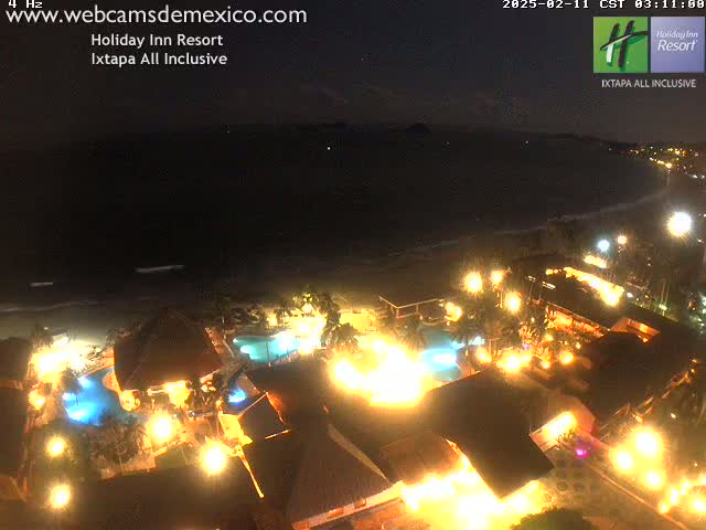 A brightly lit resort at night sits on a dark beach next to a calm, dark ocean under a clear night sky.