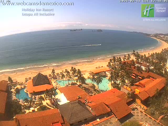 An aerial view shows a resort complex with red-tiled roofs and swimming pools next to a sandy beach and calm ocean under a sunny sky.