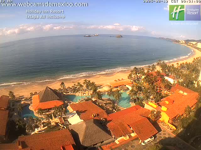 An aerial view of a resort on a sandy beach with a calm ocean under a mostly sunny sky.