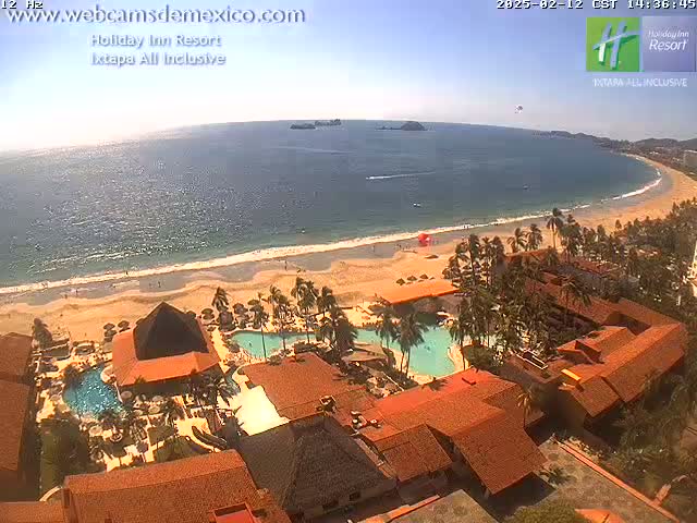 An aerial view of a resort complex on a sunny day, showing a sandy beach, ocean, and swimming pools.