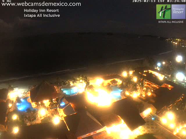 A brightly lit resort at night, situated on a beach next to a dark ocean under a clear sky.