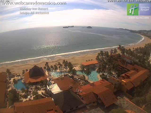 An aerial view of a resort complex with red tile roofs, multiple pools, and palm trees, situated on a sandy beach next to a calm, clear ocean under a mostly sunny sky.