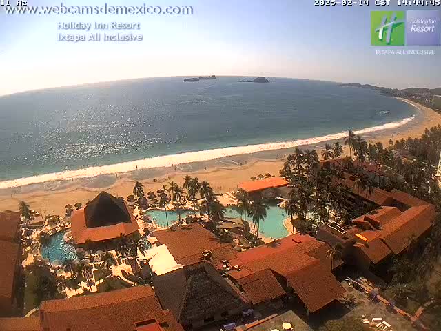 An aerial view shows a resort with multiple pools and terracotta-roofed buildings situated on a sandy beach next to a calm, sunny ocean.