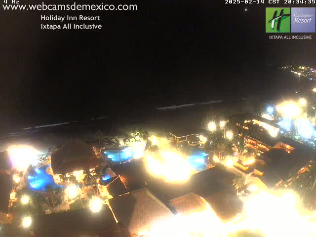 A brightly lit resort at night sits on a dark ocean shore under a clear, dark sky.