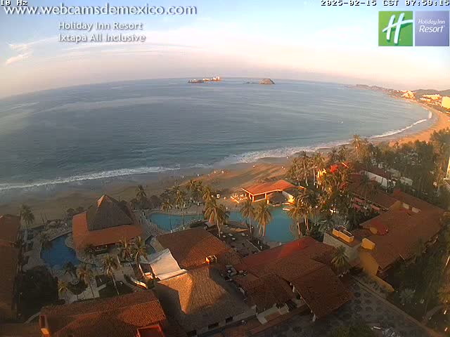 An aerial view shows a resort with red tile roofs situated on a beach next to a calm ocean under a clear, sunset sky.