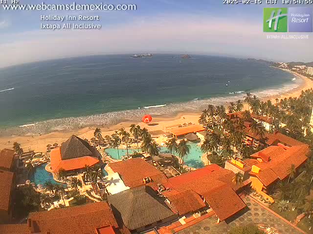 An aerial view of a resort on a sandy beach, with calm ocean waves under a partly sunny sky.
