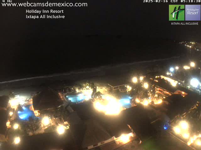 A nighttime aerial view of a brightly lit resort complex next to a dark ocean under a clear sky.