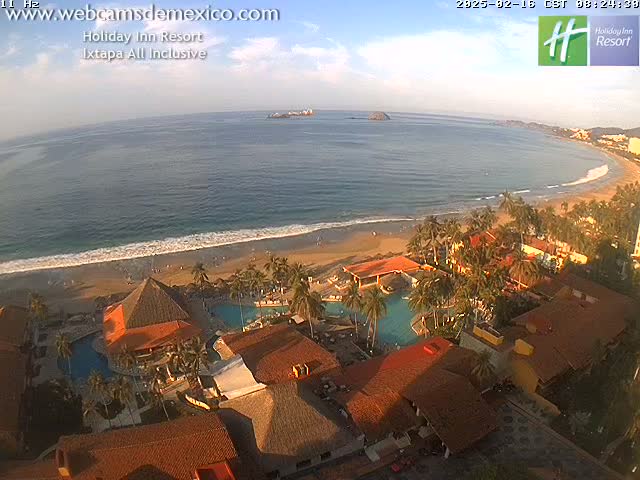 A beachfront resort with swimming pools, palm trees, and terracotta-roofed buildings is shown under a mostly clear, sunny sky.