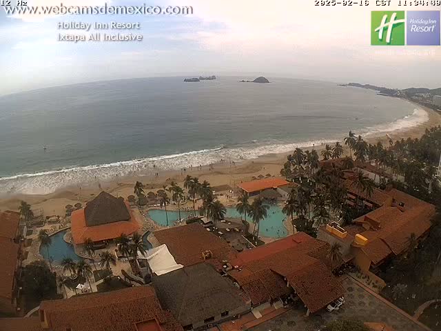 An aerial view shows a resort complex with multiple pools, surrounded by palm trees, situated on a beach with gentle waves under an overcast sky.