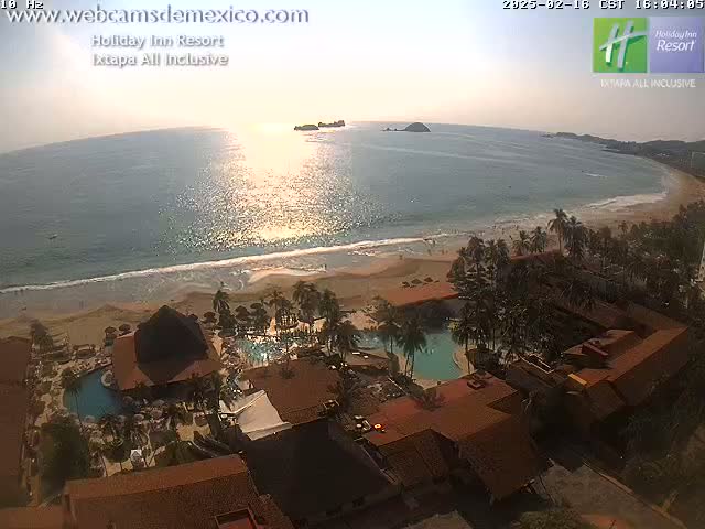 An aerial view shows a resort with swimming pools nestled among palm trees on a sandy beach, with calm ocean waters reflecting the setting sun.