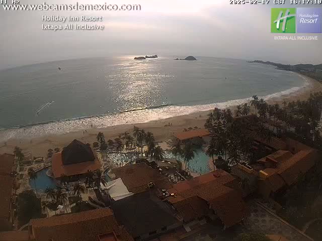 An aerial view of a resort overlooking a calm, sunlit ocean and sandy beach with palm trees.