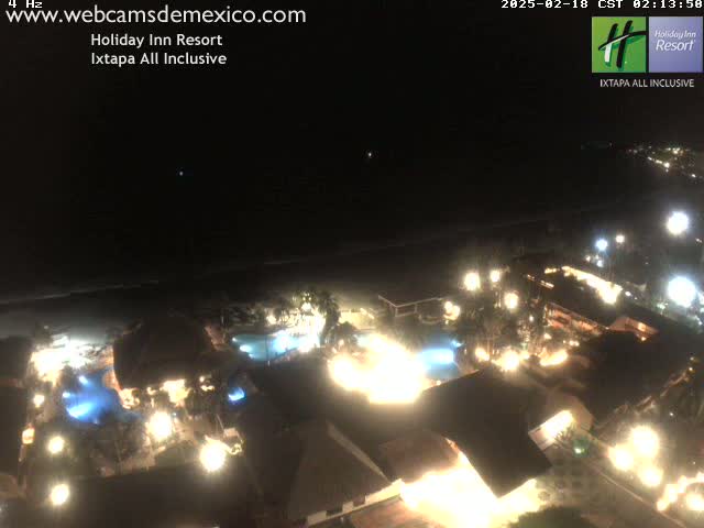 A nighttime aerial view of a brightly lit resort complex next to a dark ocean under a clear sky.