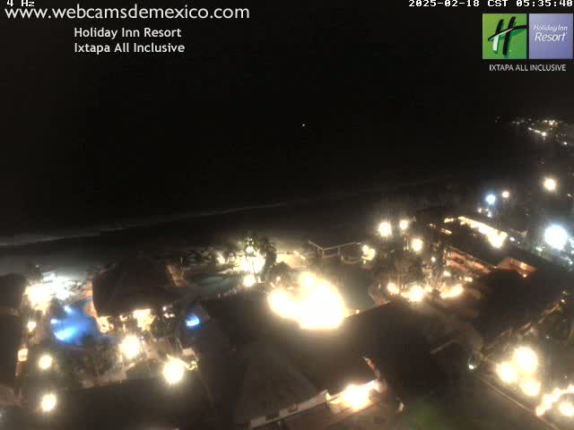 A nighttime aerial view of a brightly lit resort complex next to a dark ocean under a clear sky.