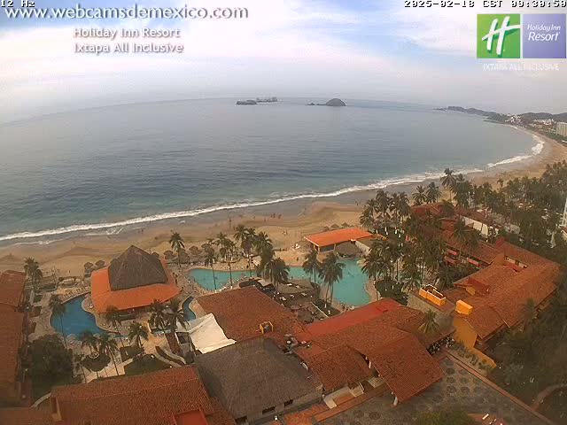 An aerial view of a resort with multiple pools and terracotta-roofed buildings situated on a sandy beach next to a calm ocean under a mostly cloudy sky.
