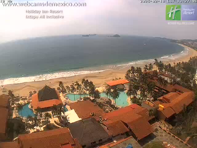 An aerial view shows a beach resort with swimming pools and terracotta-roofed buildings next to a sandy beach and ocean under hazy conditions.