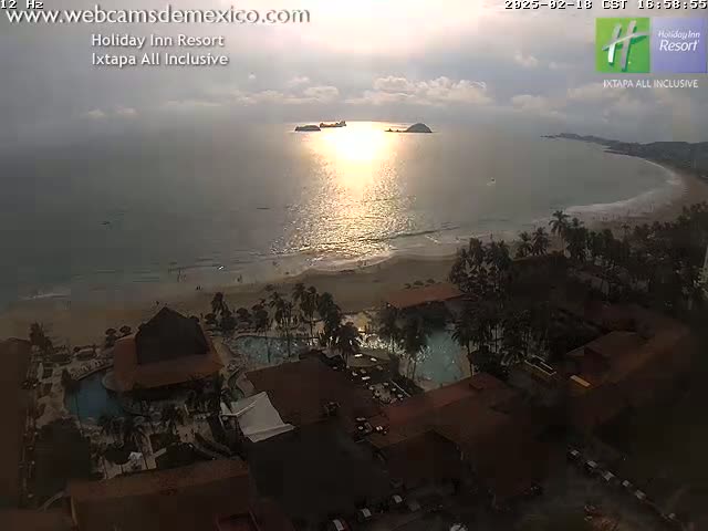 An elevated view of a resort complex with pools overlooking a sandy beach and calm ocean at sunset.
