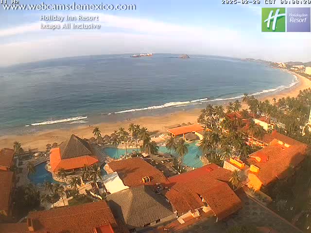 An aerial view of a resort on a sandy beach with calm ocean waves under a mostly clear sky.