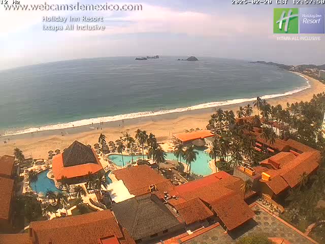 An aerial view shows a resort with red-tiled roofs and multiple swimming pools next to a sandy beach and ocean under partly cloudy skies.