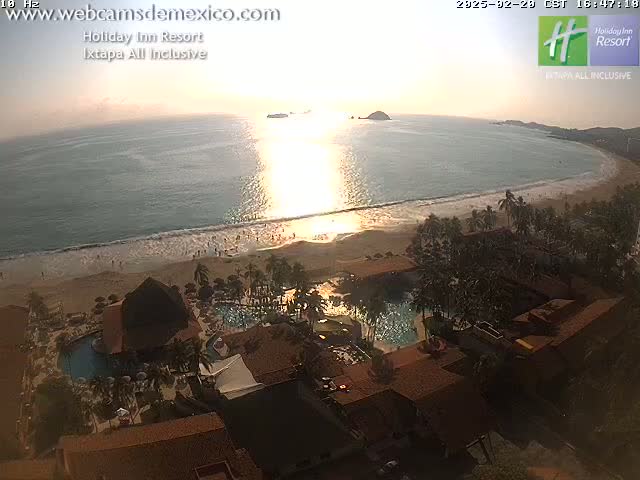 An aerial view shows a beach resort at sunset with a calm ocean reflecting the sun's golden light.