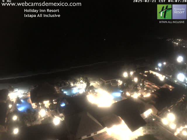 A nighttime aerial view of a resort with illuminated buildings, pools, and landscaping next to a dark ocean under a clear sky.