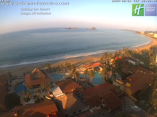 An aerial view of a beachfront resort with swimming pools, palm trees, and reddish-brown buildings, on a sunny day with calm ocean waves.