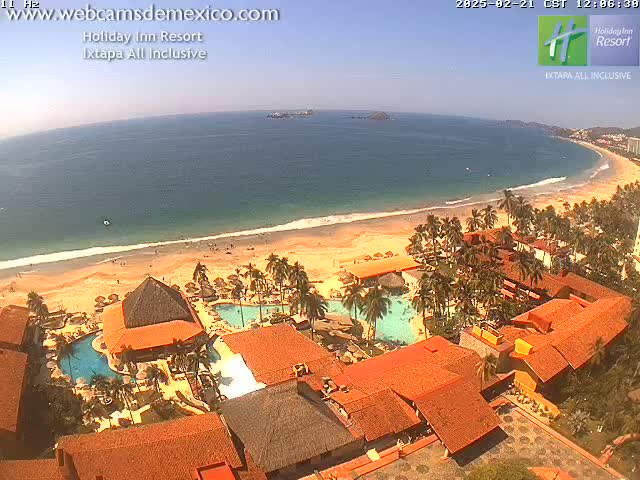A beach resort with terracotta-roofed buildings, palm trees, and swimming pools sits on a sandy beach next to a calm, blue ocean under a sunny sky.