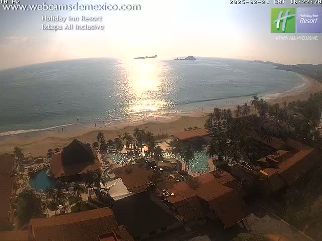 A resort with pools and palm trees sits on a sandy beach next to a calm ocean reflecting the setting sun.