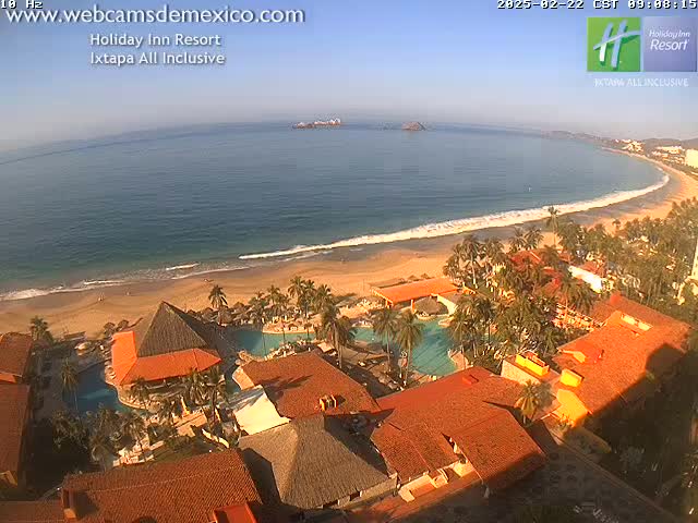 An aerial view shows a resort complex nestled beside a sandy beach and calm ocean under a clear, sunny sky.