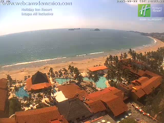 An aerial view of a beachfront resort with multiple pools and reddish-brown buildings on a sunny day with calm ocean waves.