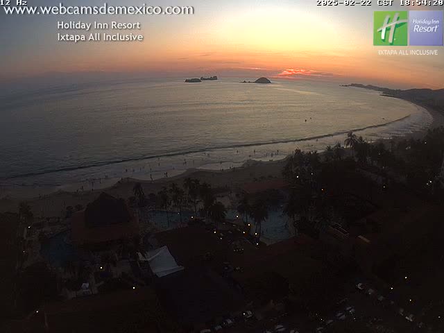 An aerial view at sunset shows a resort complex next to a beach with people on it and islands in the distance.