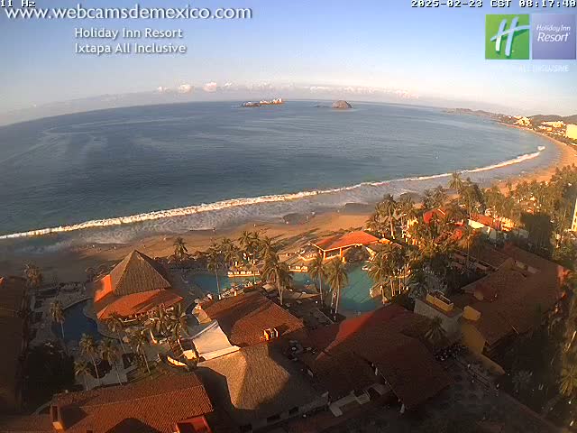 An aerial view of a resort complex with red-tiled roofs situated near a sandy beach and ocean under a clear, sunny sky.