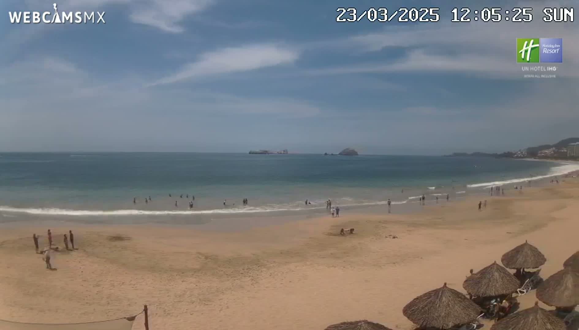 A sandy beach with people swimming and relaxing under thatched umbrellas, on a partly sunny day, with calm ocean waves and distant islands visible.