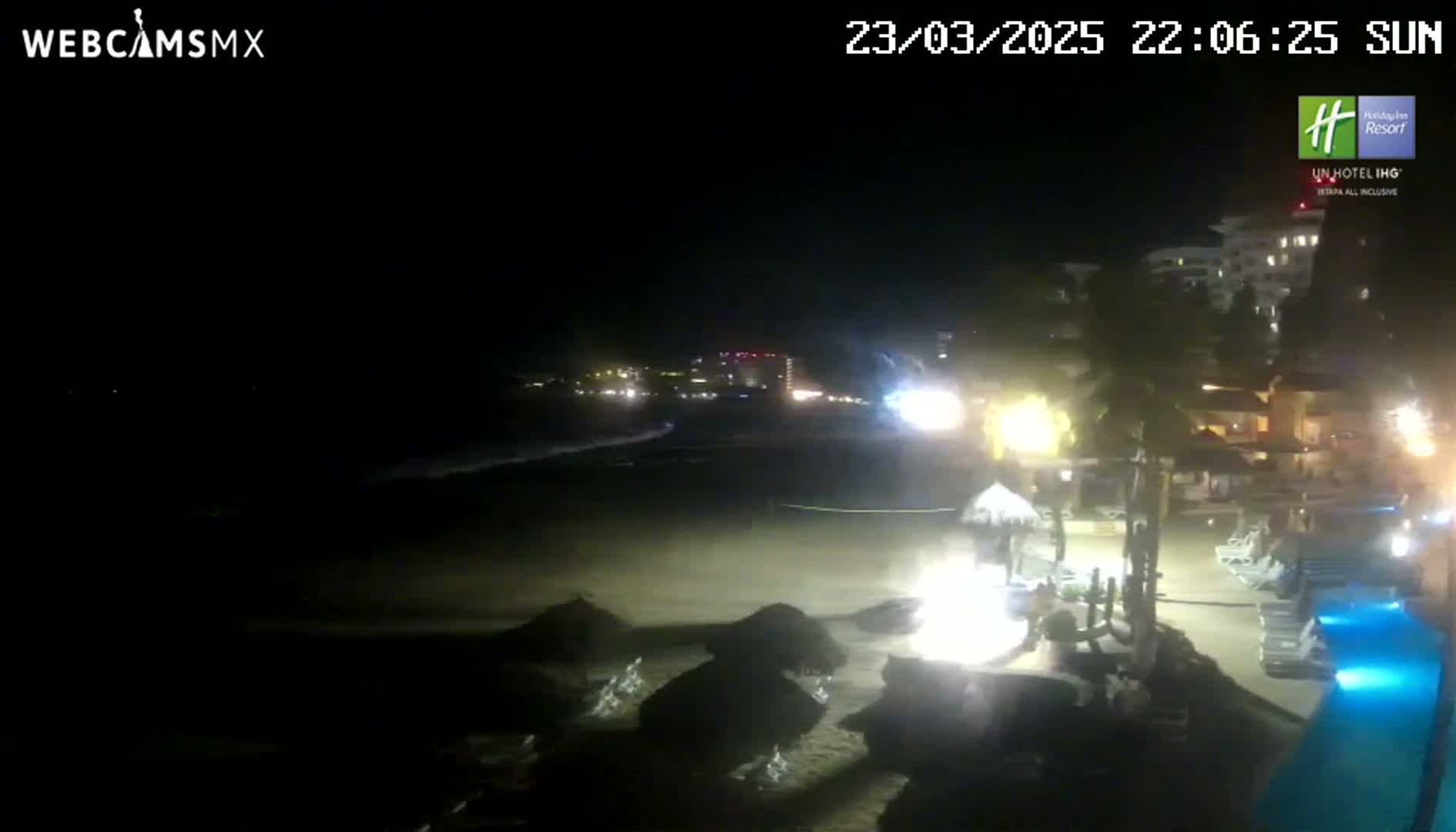 A nighttime beach scene shows a dark ocean with waves, a sandy shore with beach umbrellas, a resort with lights, and a lit pool.