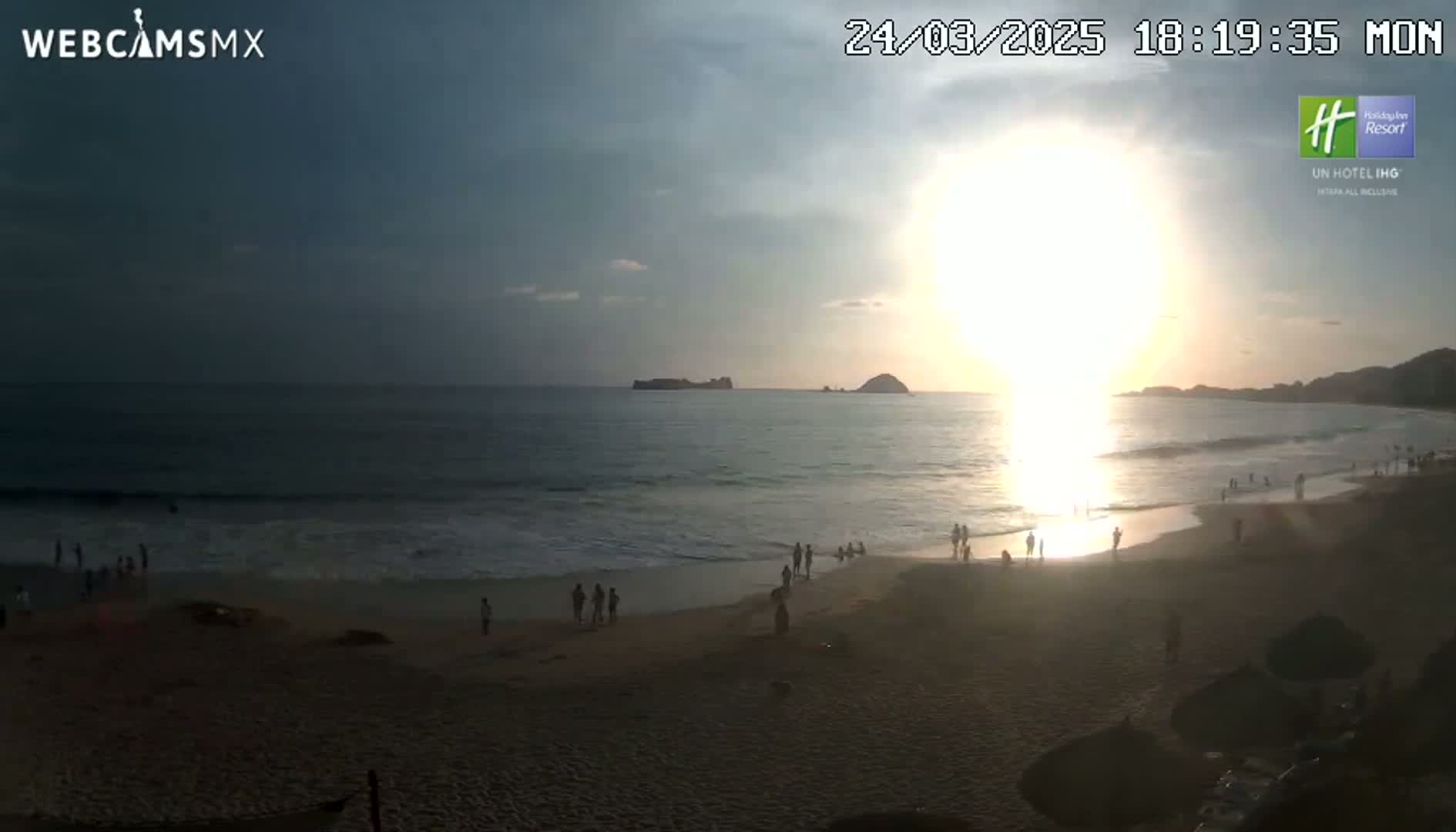 A beach scene at sunset shows people on the sand and in the water, with small islands visible in the distance under a partly cloudy sky.