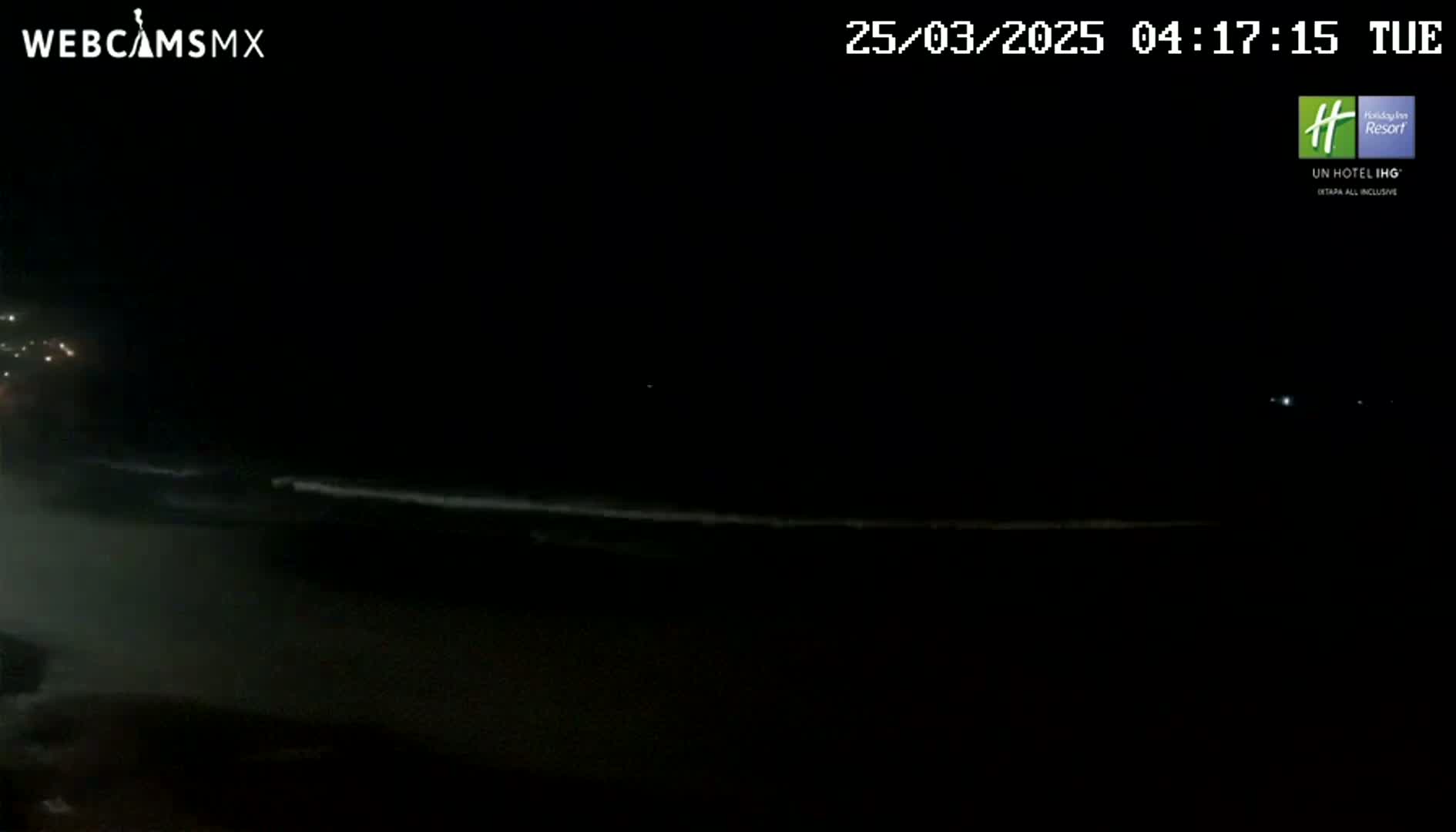 A dark, nighttime beach scene shows a faint line of waves breaking on the shore under a clear, starless sky.