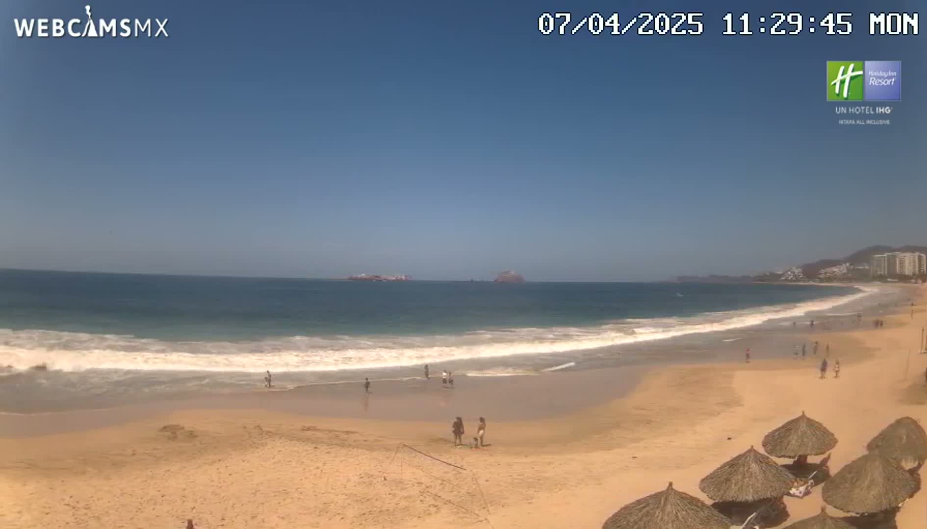 A sandy beach with calm waves and a few people on it stretches out under a clear blue sky.