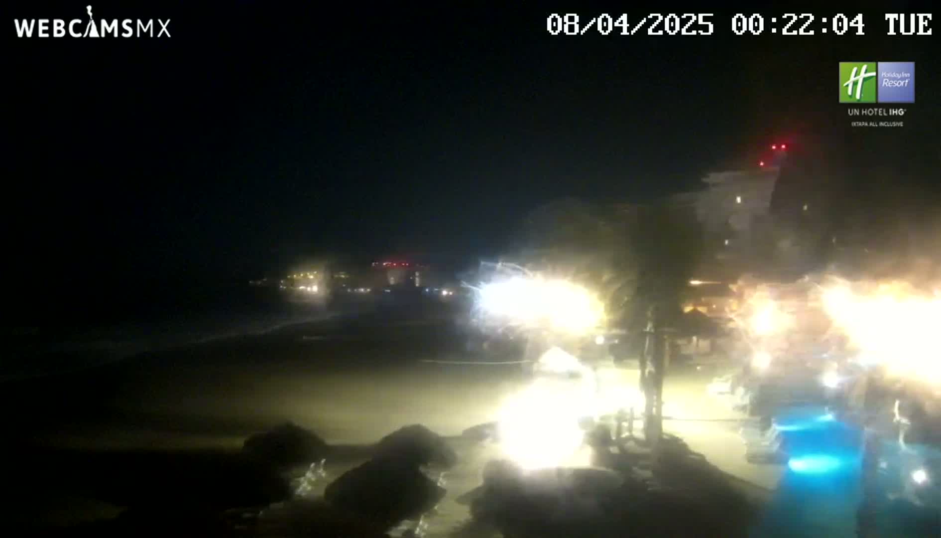 A nighttime beach scene shows a dark ocean, a sandy shore, brightly lit buildings and palm trees, and a pool with illuminated blue water.