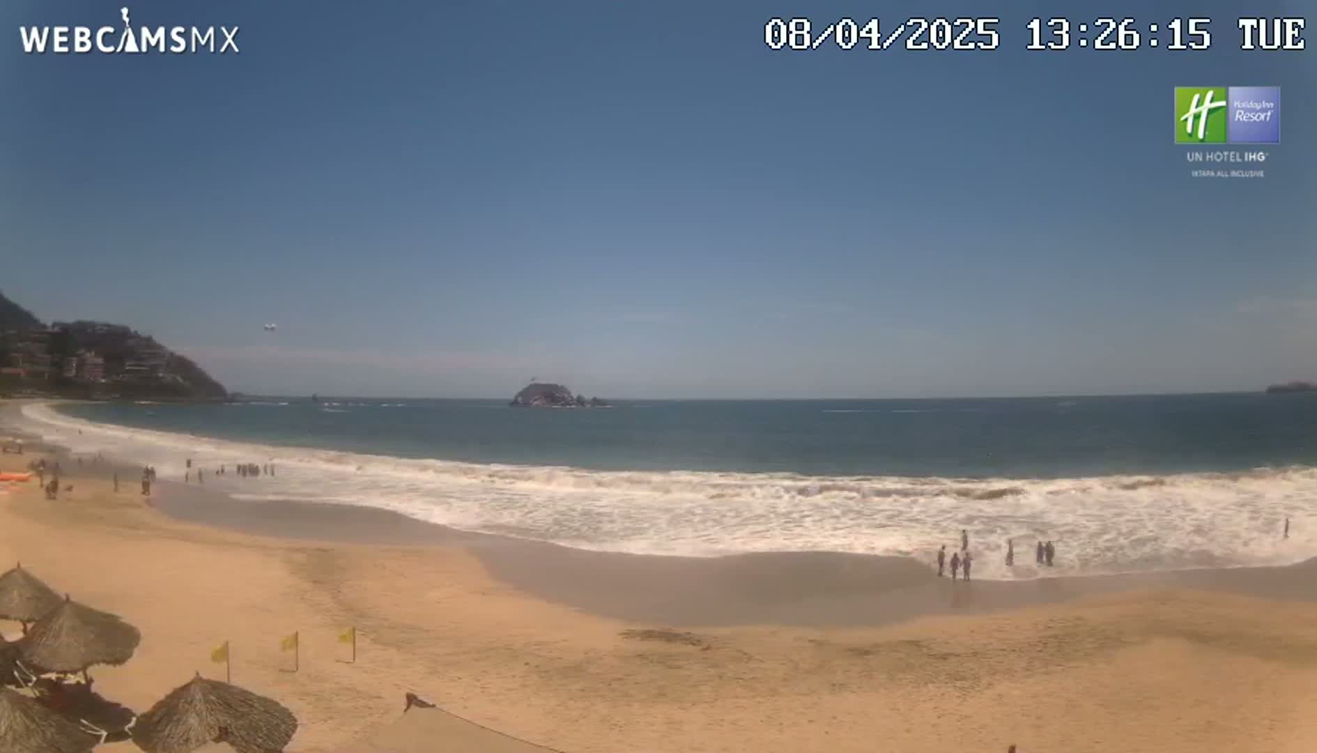 A sandy beach with calm waves under a clear blue sky, people are seen enjoying the beach.