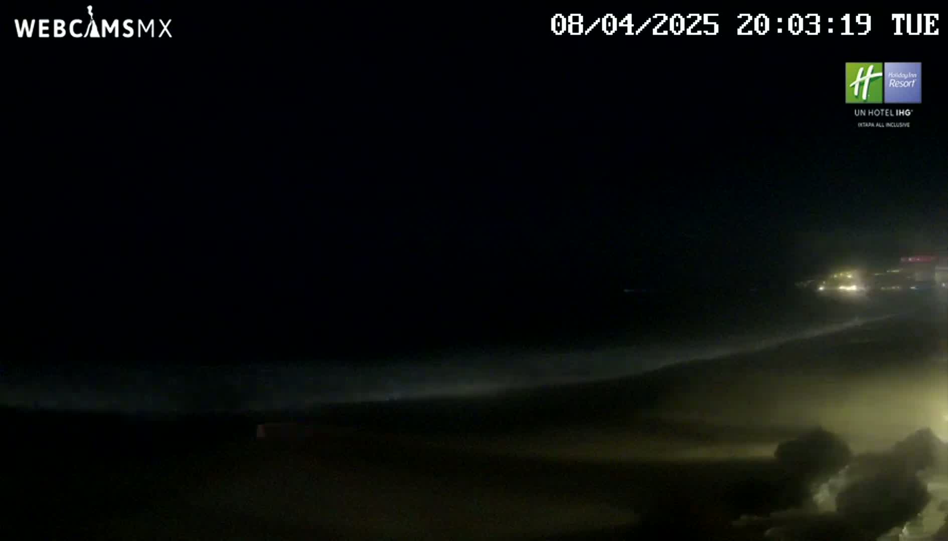 A dark nighttime beach scene shows waves crashing on the shore under a mostly clear sky.
